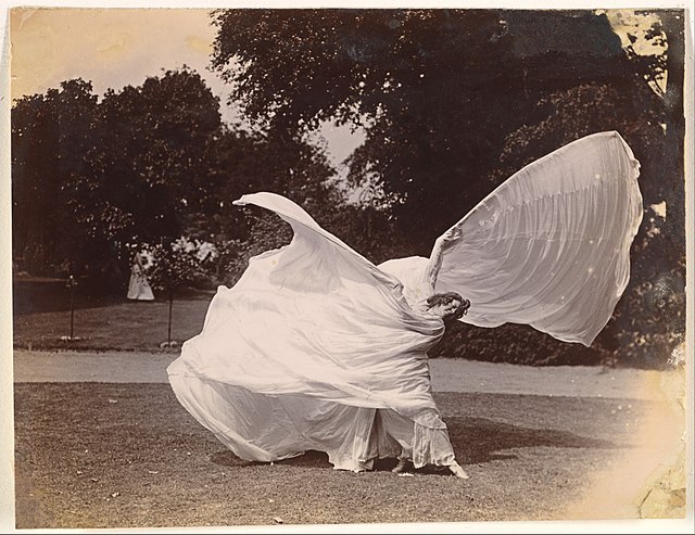 Eine Schwarz-Weiß-Fotografie; Loie Fuller tanzt in einem hellen Gewand im Park