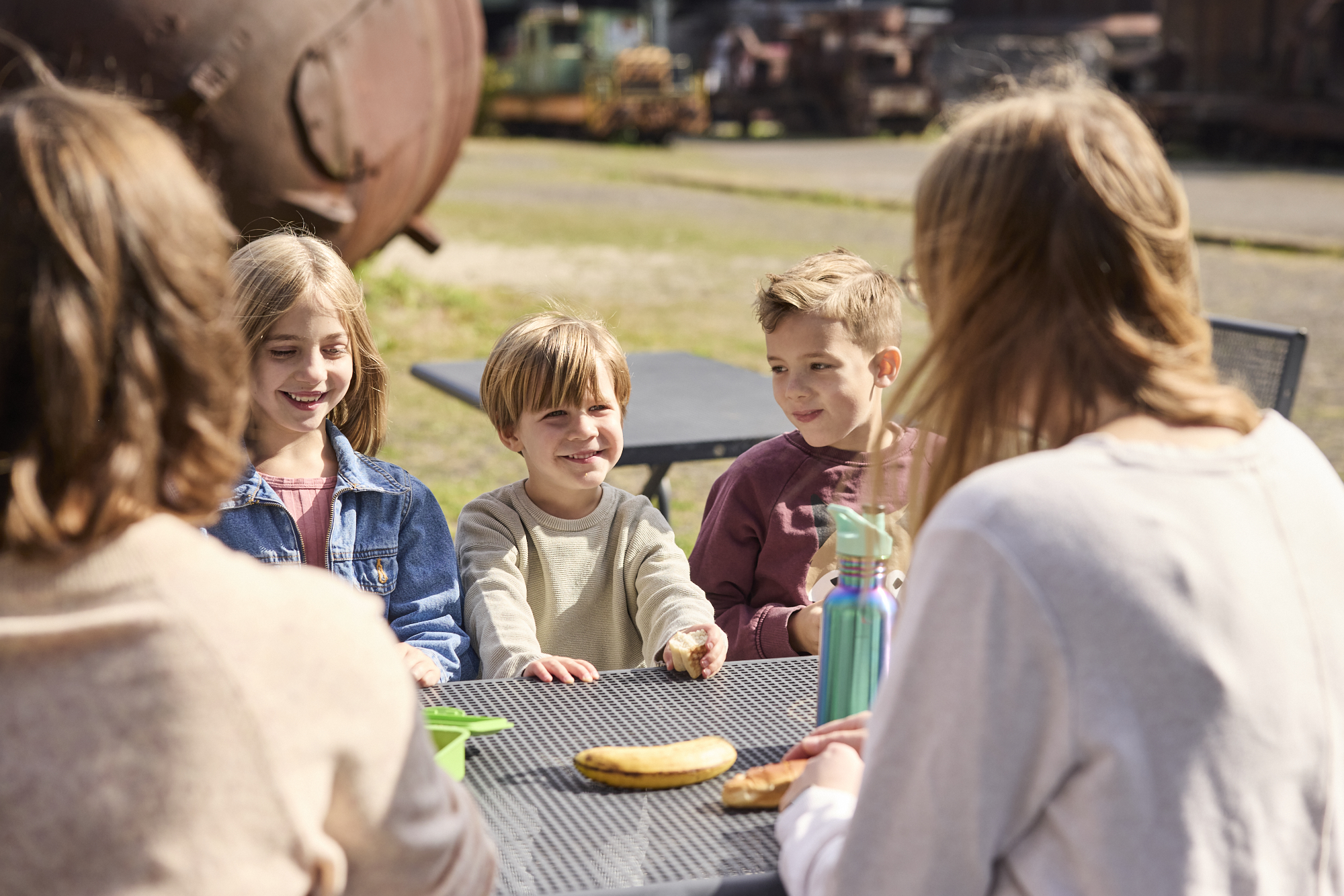 3 Kinder sitzen an einem Tisch und vor Ihnen liegt Essen. Im Hintergrund erkennt man das Geländer der Henrichshütte