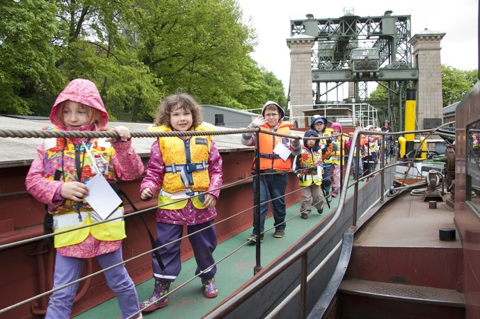 Kinder mit Westen laufen über das Deck eines Schiffes. Im Hintergrund das Schiffshebewerk.
