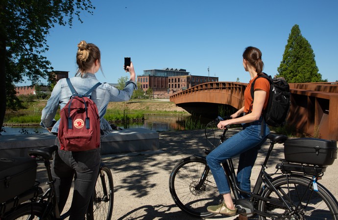 Zwei Fahradfahrerinnen stehen vor einer Brücke und machen ein Foto von der Spinnerei des Textilwerks.