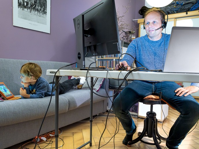 Ein Mann sitzt in seinem Wohnzimmer am Schreibtisch, neben ihm ein Kind auf einem Sofa an einem Tablet