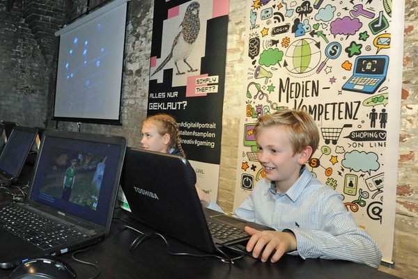 Zwei Kinder sitzen an Computern. Im Hintergrund ein Banner mit Zeichnungen und der Aufschrift "Medienkompetenz".
