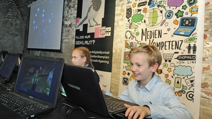 2 Kinder sitzen an Computern, im Hintergrund an der Wand Banner mit Zeichnungen und der Aufschrift "Medienkompetenz"