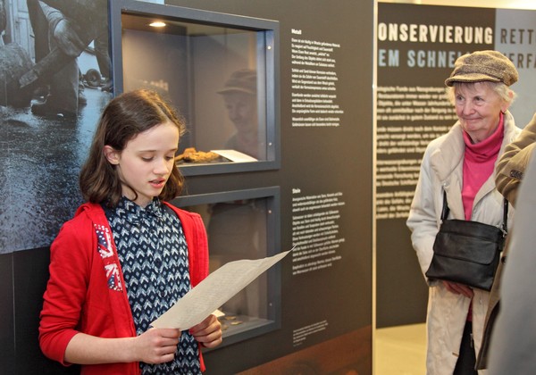 Ein Mädchen liest in einer Ausstellung einen Text von einem Blatt ab, rechts im Bild eine Seniorin.