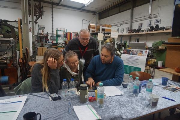 Vier Personen sitzen um ein Tablet und schauen sich konzentriert verschiedene Inhalte an.