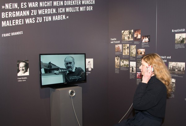 Eine Frau mit Kopfhörer verfolgt in einer Ausstellung ein Videointerview mit einem Zeitzeugen. An der Wand sind Fotos und Texte zu sehen.